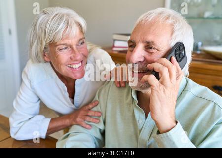 Senior couple enjoying retirement, with the man talking on a smartphone, possibly with a financial advisor, while the woman smiles happily beside him. Stock Photo
