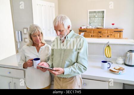 A senior couple is actively engaged in digital estate planning and financial protection, reviewing important documents on a tablet in their modern kit Stock Photo