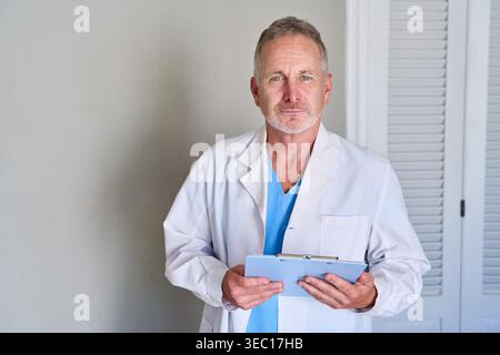 A senior male doctor or medical professional stands confidently, holding a clipboard. He is dressed in a white lab coat over blue scrubs, representing Stock Photo