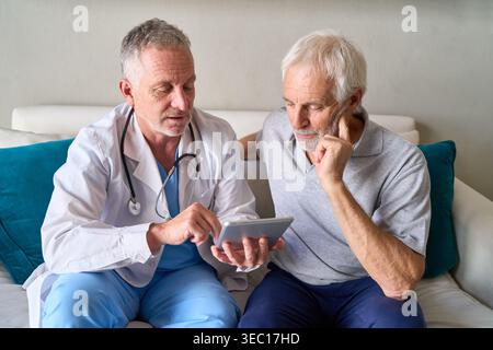 Telemedicine consultation for seniors — a male doctor in a white coat and stethoscope is explaining digital health records on a tablet to an attentive Stock Photo
