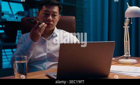 Serious Asian 40s business man recording audio message talk mobile phone at night evening office Korean businessman record voice mail talking to Stock Photo