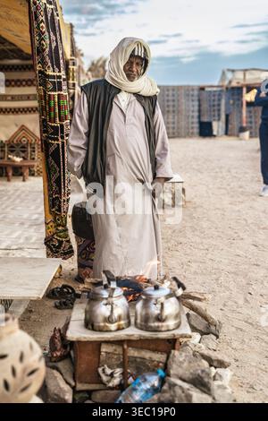 Marsa alam, egypt 21 april 2026: bedouin man in traditional galabeya and headscarf brewing tea over a campfire in the marsa alam desert, authentic cul Stock Photo