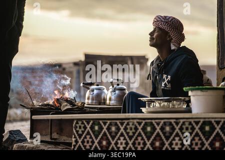 Marsa alam, egypt 21 april 2026: young bedouin man wearing a keffiyeh smiling, preparing tea in kettles over a campfire at sunset in the marsa alam de Stock Photo