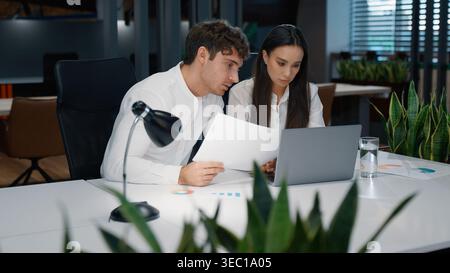 Two business partners talking looking in papers with stats data graphs of project marketing result reading documents computer work in office Stock Photo