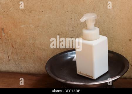 White plastic pump dispenser bottle for liquid soap resting calmly on dark metal dish against textured beige wall creating soothing and minimalist bat Stock Photo