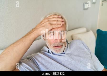 Health issues in older age — a senior man with a white beard and gray hair is shown with his eyes closed, holding his hand to his forehead, indicating Stock Photo