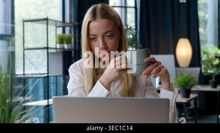Caucasian businesswoman drinking coffee in morning office typing laptop work web job internet using business woman drink tea holding cup female girl Stock Photo