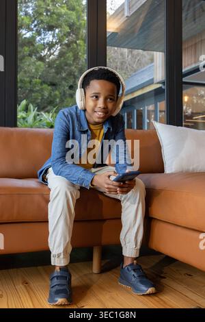 African American preteen boy sitting in living room on tan sofa wearing headset and holding phone Stock Photo
