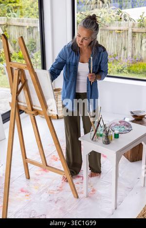 Senior female artist painting at easel in home studio with palette and brushes on table Stock Photo