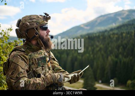 Bearded Soldier In Tactical Gear Using Tablet With Radio Headset And Mountain Landscape Background Stock Photo