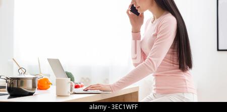 Woman Working From Home in Modern Kitchen While Talking on the Phone and Using a Laptop Stock Photo