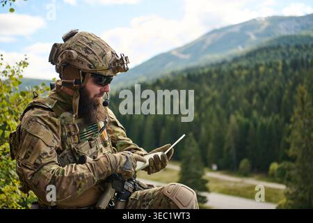 Tactical Soldier Using Tablet and Radio Communications While On Patrol in Forested Mountain Terrain Stock Photo