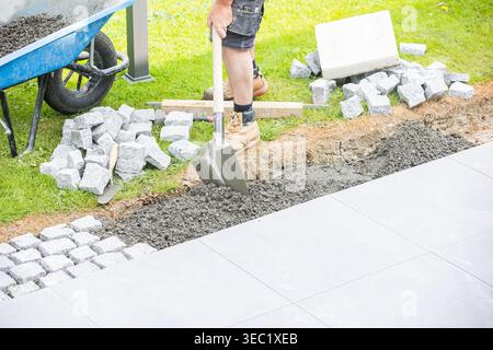 Construction worker using a shovel to prepare the base for cobblestone pavement installation, with tools and materials visible on a landscaping projec Stock Photo
