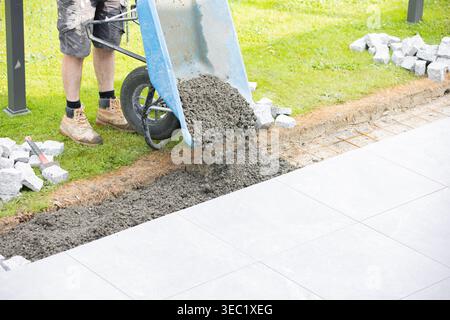 Construction worker pouring fresh concrete from a wheelbarrow to prepare the base for cobblestone pavement installation on a landscaping project. Stock Photo