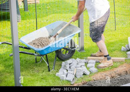 Construction worker using a shovel to prepare the base for cobblestone pavement installation, with tools and materials visible on a landscaping projec Stock Photo