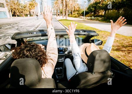Young adult couple raising hands while riding in convertible car at city Stock Photo