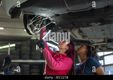 Two women car mechanics using a torch and a digital tablet while checking the undercarriage of a vehicle in a busy repair shop. Stock Photo