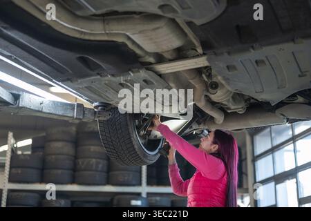 Focused professional female auto mechanic using a flashlight to inspect a jacked-up car, brake system checks at a repair shop. Stock Photo