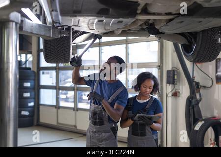 Two African American mechanics, a man and a woman, inspecting car undercarriage with a torch and tablet for routine checkups. Stock Photo