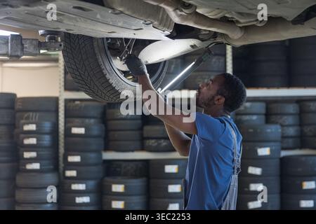 African American auto mechanic inspecting the underside of a car using a torch at a professional repair service with stocked tyres and tools. Stock Photo