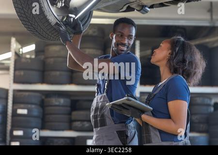 Diverse pair of smiling mechanics working together, inspecting the underside of a lifted vehicle in a modern repair shop. Stock Photo