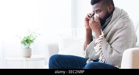 Man Wrapped in Blanket Sitting on Couch, Feeling Unwell and Using Tissue Indoors Stock Photo