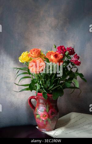 Vibrant orange and yellow roses in a rustic red ceramic pitcher, set against a textured grey backdrop with a vintage letter Stock Photo
