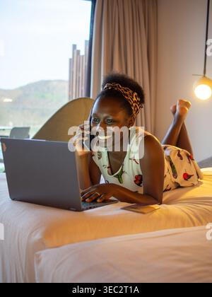 Happy African woman lying on bed, using laptop with a smile in a bright bedroom. Stock Photo