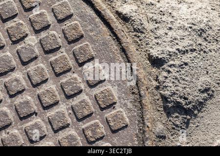 Abstract urban photo, background texture of a rusty cast-iron manhole cover set into rough concrete and sand, highlighting a geometric grid pattern, w Stock Photo