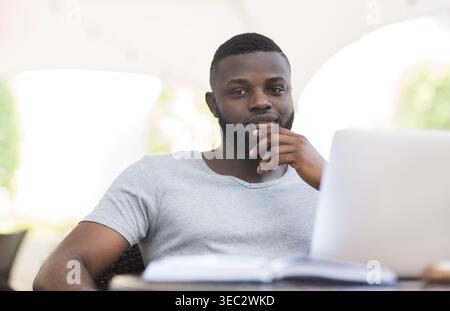 Young Man Working Thoughtfully on Laptop at Outdoor Cafe During a Sunny Day Stock Photo