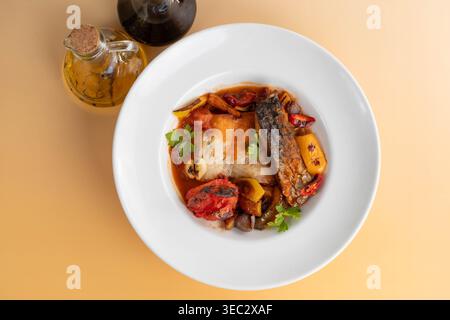 Top view flat lay of baked carp fish fillet in tomato sauce with roasted vegetables, served in a deep white plate next to oil and vinegar cruets Stock Photo