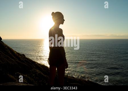 A young woman in sportswear stands against the backdrop of the sun setting over the ocean. She enjoys the peaceful moment on a coastal rock, embracing Stock Photo
