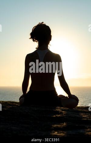 A young woman sits in a meditative pose on a rock, facing the sea as the sun rises. The warm light creates a serene atmosphere in a beautiful shorelin Stock Photo