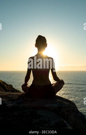 A young woman sits in a meditative pose on a rock, facing the sea as the sun rises. The warm light creates a serene atmosphere in a beautiful shorelin Stock Photo
