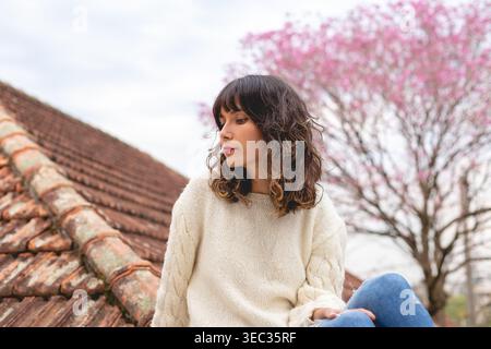 A young woman sits on a rooftop wearing a light sweater. She looks thoughtful as she gazes away from the camera. A pink flowering tree is visible in t Stock Photo