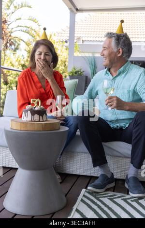 Couple celebrating on patio wicker sofa wearing gold party hats holding wine near chocolate 50 cake Stock Photo
