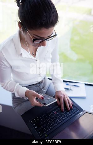 Middle Eastern businesswoman multitasking on smartphone and laptop in modern open office during meeting Stock Photo