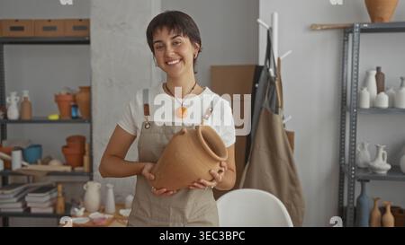Young woman holding clay pot with hands, smiling and tilting head in a pottery studio; serenity craft. Stock Photo