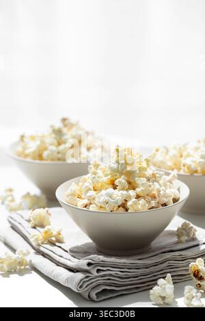 Modern lifestyle composition featuring several white ceramic bowles filled with freshly popped popcorn, placed on a folded cloth atop a white wooden t Stock Photo