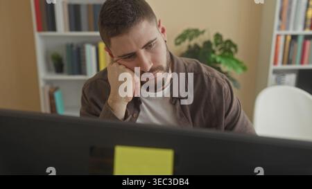 Young hispanic man with beard resting head on fist at computer desk, looking down, bookshelf and plant behind him in a building; pensive reflection. Stock Photo