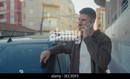 Man holding smartphone to ear while leaning on car door on street, smiling slightly and resting elbow on roof; casual conversation thoughtful. Stock Photo