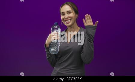 Woman holding a large plastic water bottle and waving right hand in studio with purple backdrop, smiling at camera; hydration joy. Stock Photo