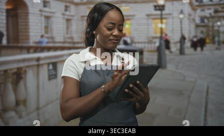 Woman in apron taps a tablet while holding it, smiling and checking orders on a cobblestone street outside a stone building; quiet focus service. Stock Photo