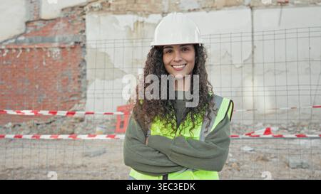 Woman architect wearing hardhat and reflective vest, arms crossed at building site; confidence leadership professionalism. Stock Photo