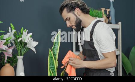 Young hispanic man florist wearing apron carefully wipes a snake plant leaf with an orange cloth beside lilies and ceramic vases in a floral studio; s Stock Photo