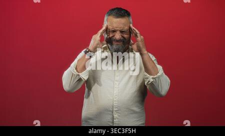 Man pressing temples with hands, bearded middle age man wearing a white shirt in studio against a red wall; pain. Stock Photo