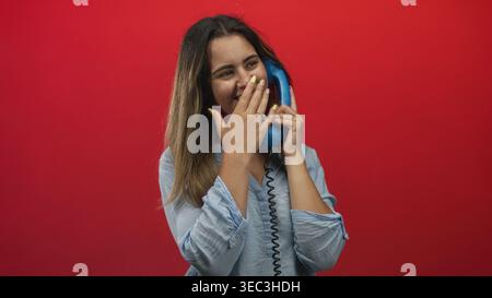 Woman holding blue telephone handset with hand over mouth laughing in red studio wearing light blue shirt and yellow nails; playful joy. Stock Photo