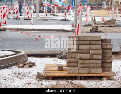A wooden pallet stacked with concrete paving blocks stands at a road construction site partially covered with snow. The area is secured with red?and?w Stock Photo