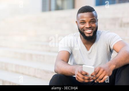 Young Man Smiling While Sitting on Steps and Using Smartphone Outdoors in Bright Sunlight Stock Photo