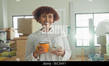 Woman holds smartphone and orange mug, hand tapping phone while surrounded by moving boxes in building; settling in contentment. Stock Photo